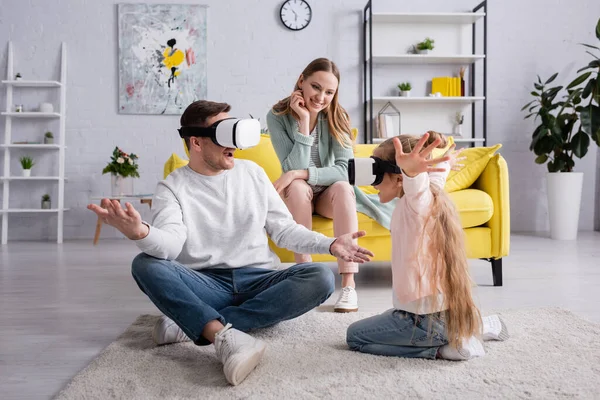 Padre e hija jugando en auriculares de realidad virtual cerca de la madre en el sofá - foto de stock