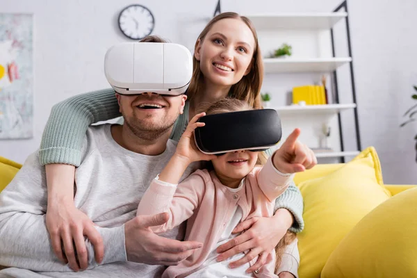 Mujer sonriente abrazando a su hija y marido en auriculares vr en el sofá - foto de stock