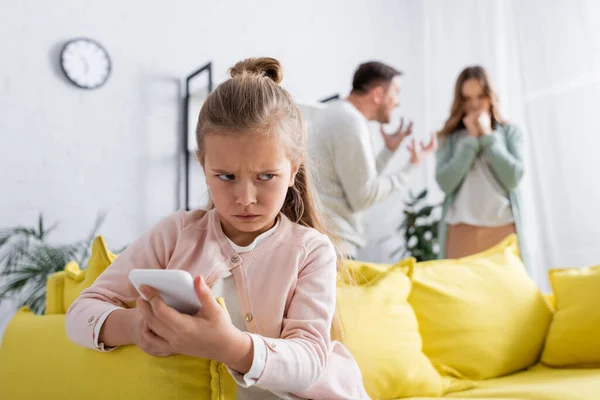 Trauriges Kind nutzt Smartphone, während sich Eltern vor verschwommenem Hintergrund streiten — Stockfoto