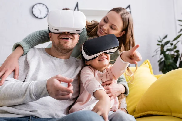 Lächelndes Mädchen zeigt mit dem Finger beim Spielen in vr Headset in der Nähe der Eltern — Stockfoto