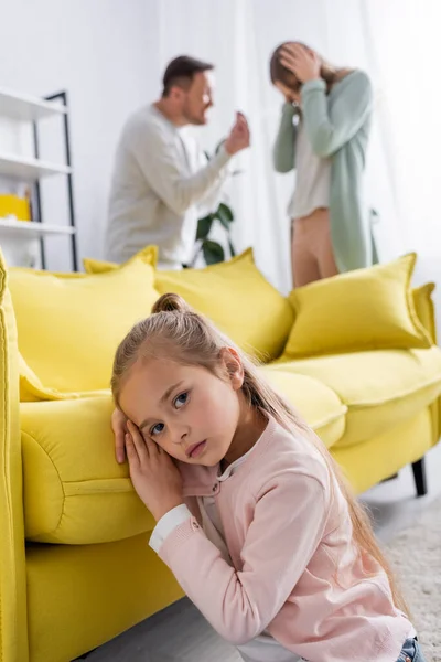 Chica mirando a la cámara cerca del sofá y los padres que tienen conflicto sobre fondo borroso - foto de stock