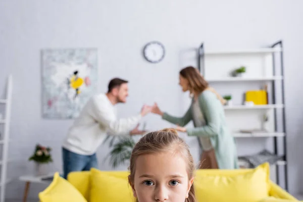 Girl looking at camera with blurred parents quarrelling at background at home — Stock Photo