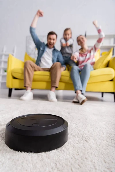 Robotic vacuum cleaner on carpet near family on couch on blurred background — Stock Photo