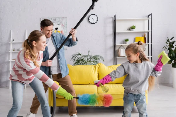 Chica en guantes de goma jugando con los padres sosteniendo suministros de limpieza en casa - foto de stock