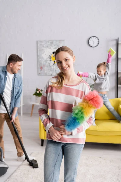 Donna sorridente tenendo spazzola polvere vicino alla famiglia su sfondo sfocato — Foto stock