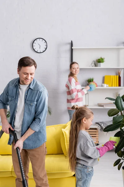 Hombre sonriente usando aspiradora cerca de la planta de limpieza hija - foto de stock