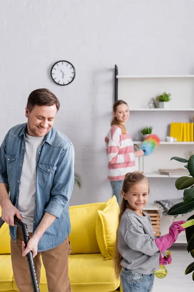 Sorridente padre in possesso di aspirapolvere vicino figlia impianto di pulizia e moglie a casa — Foto stock