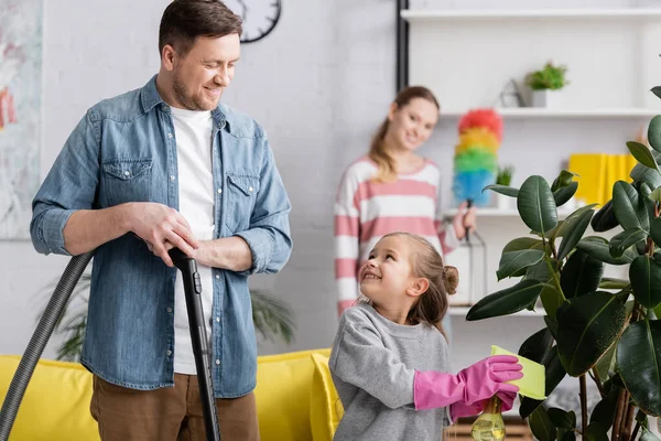 Chica planta de limpieza y mirando a padre con aspiradora en casa - foto de stock