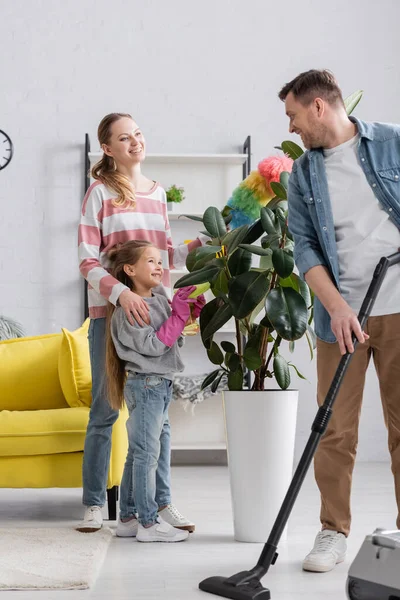 Sorrindo homem com aspirador de pó olhando para a família perto de planta em casa — Fotografia de Stock