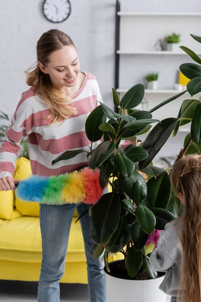 Mujer sonriente planta de limpieza con cepillo de polvo cerca de la hija - foto de stock