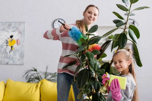 Chica sonriente sosteniendo detergente y trapo cerca de la madre y la planta - foto de stock