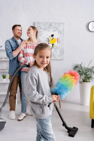 Enfant avec brosse à poussière regardant la caméra près des parents et aspirateur — Photo de stock