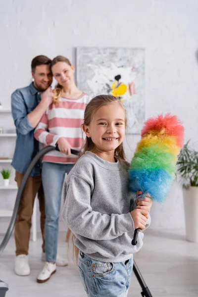 Ragazzo positivo con spazzola antipolvere vicino ai genitori con aspirapolvere su sfondo sfocato — Foto stock