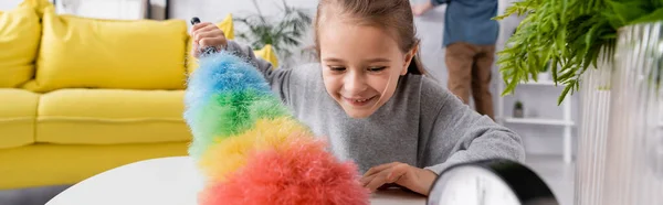 Chica sonriente usando cepillo de polvo en la mesa en casa, pancarta - foto de stock