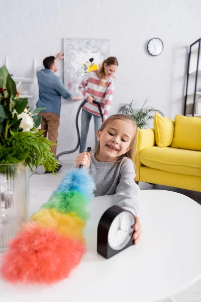 Enfant tenant une table de brosse à poussière pendant que les parents nettoient le salon — Photo de stock