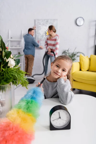 Enfant souriant avec brosse à poussière regardant la caméra près de l'horloge et les plantes sur la table — Photo de stock