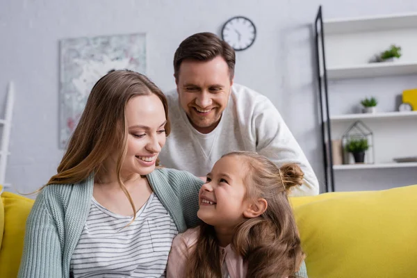 Mujer sonriente mirando a su hija cerca de marido borroso en la sala de estar - foto de stock