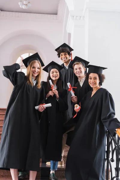 Graduados multiculturales sonrientes en vestidos académicos con diplomas - foto de stock