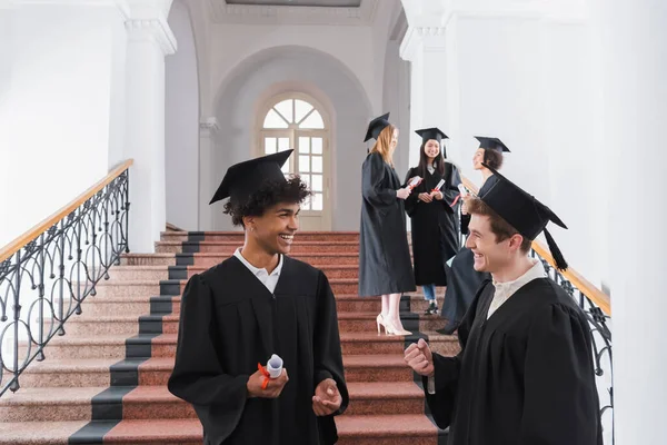 Sonriente afroamericano graduado con diploma de pie cerca de amigo mostrando sí gesto en la universidad - foto de stock