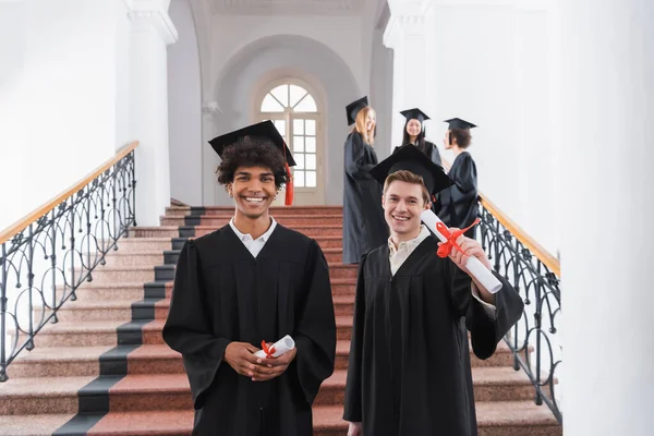 Positive interracial students holding diplomas in university — Stock Photo