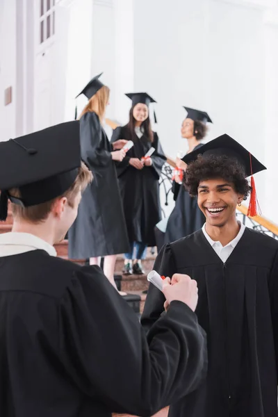 Smiling african american graduate with diploma standing near friend on blurred foreground — Stock Photo