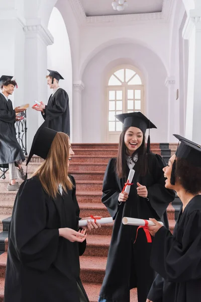 Estudiantes multiétnicos sonrientes con diplomas hablando en la universidad - foto de stock