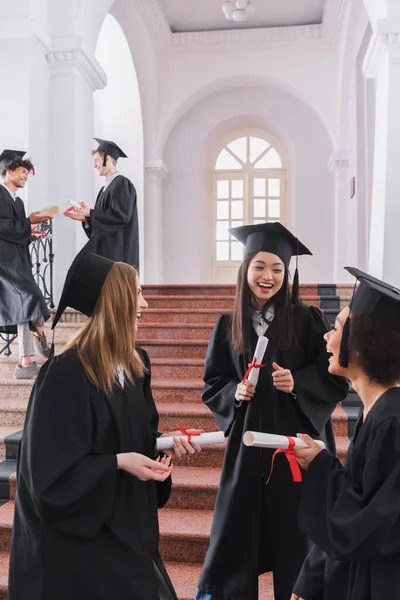 Smiling multiethnic students with diplomas talking in university — Stock Photo
