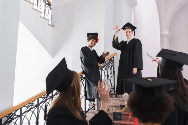 Graduados multiétnicos sonrientes con diplomas saludando a sus amigos en un primer plano borroso - foto de stock