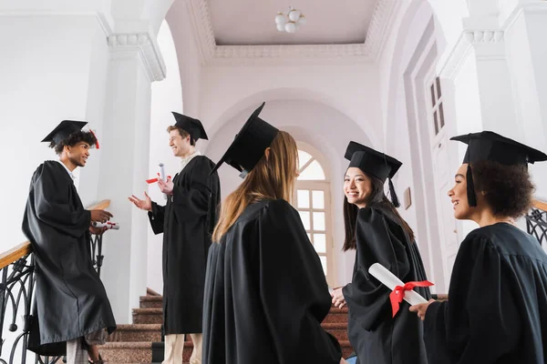 Graduados interraciales en vestidos académicos y gorras hablando en la universidad - foto de stock