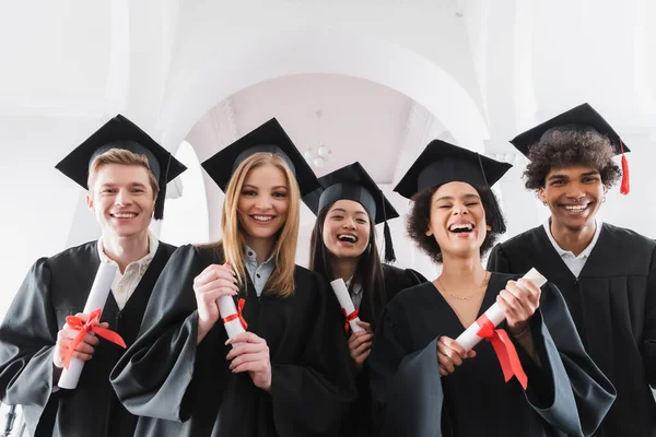 Estudiantes multiculturales positivos con diplomas sonriendo a la cámara - foto de stock