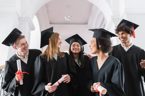 Улыбающиеся межрасовые выпускники смотрят друг на друга в университете — стоковое фото