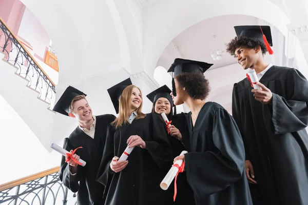 Vista basso angolo di sorridente laureati interrazziale in piedi in università — Foto stock