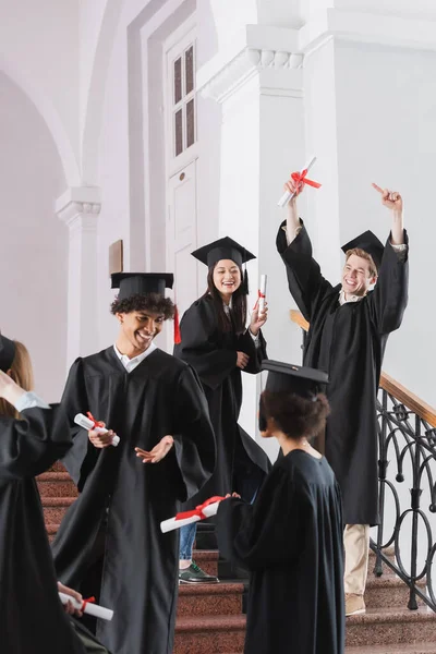 Cheerful student pointing at diploma near multicultural friends on blurred foreground — Stock Photo