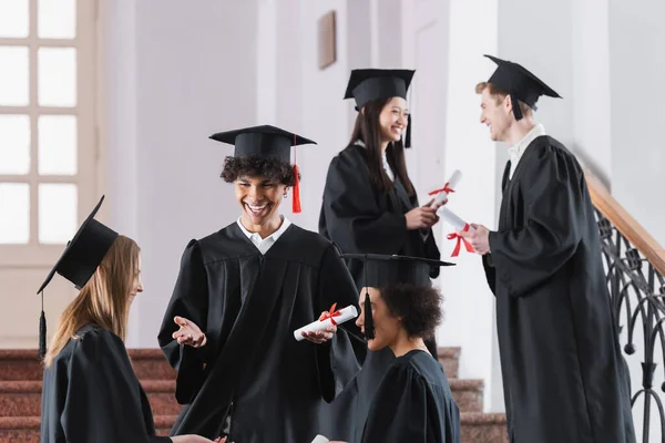 Positive african american graduate in cap talking to friends in university — Stock Photo