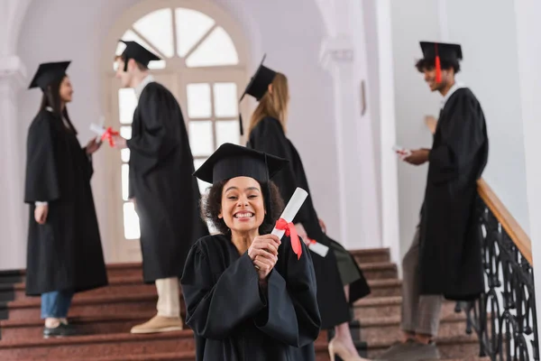 Feliz pós-graduação afro-americana segurando diploma perto de amigos turvos na universidade — Fotografia de Stock