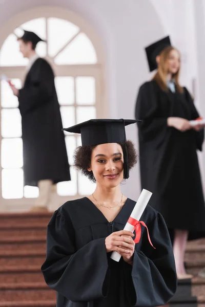 Graduado afroamericano con diploma y mirando a la cámara en la universidad - foto de stock