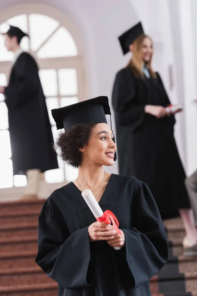 Alegre estudiante afroamericano con diploma en la universidad durante la graduación - foto de stock