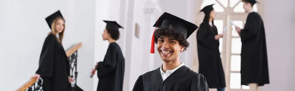 Feliz estudiante afroamericano en gorra mirando a la cámara durante la graduación, pancarta - foto de stock