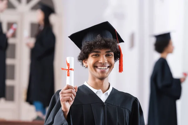 Alegre estudiante afroamericano en vestido académico y gorra con diploma - foto de stock