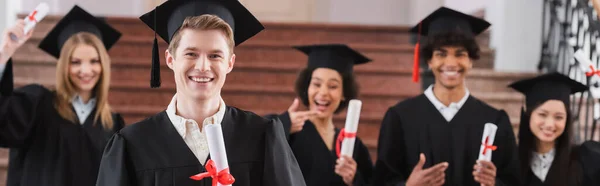 Smiling student holding diploma near multiethnic friends on blurred background, banner — Stock Photo