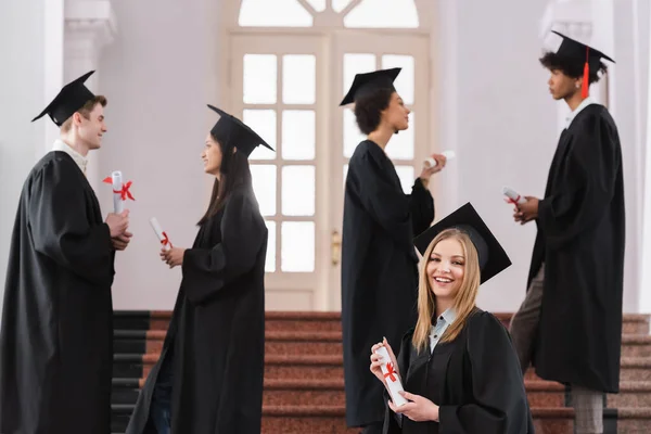 Lächelnder Junggeselle mit Diplom blickt in die Kamera in der Nähe multiethnischer Freunde — Stockfoto