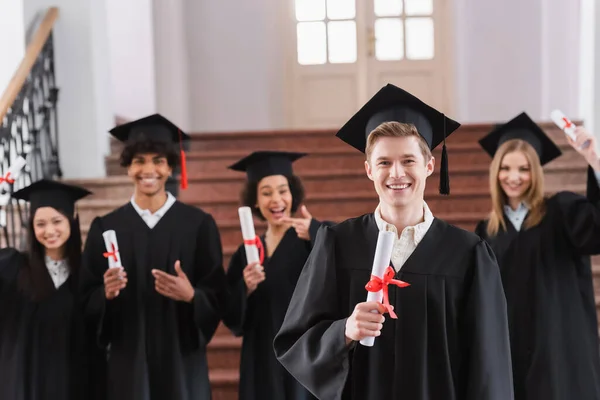 Sonriente estudiante en la celebración de la tapa diploma cerca de amigos interracial en fondo borroso - foto de stock