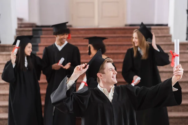 Bachelor sorridente mentre guardando il diploma vicino amici interrazziale su sfondo sfocato — Foto stock