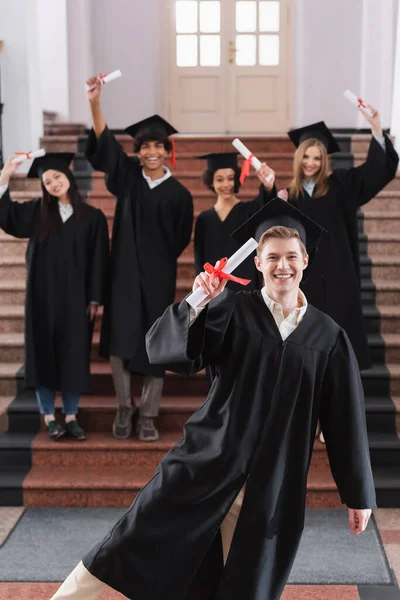 Feliz graduado sosteniendo diploma cerca de amigos interracial en vestidos académicos - foto de stock