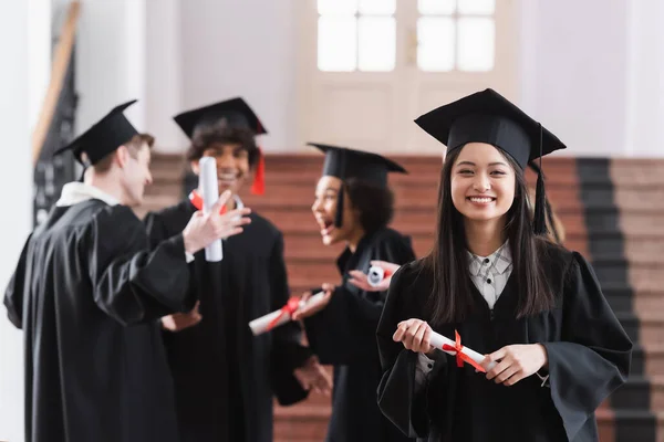 Asiático solteiro sorrindo para a câmera perto de amigos turvos na universidade — Fotografia de Stock