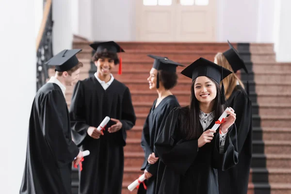 Glücklicher asiatischer Absolvent mit Diplom in der Nähe multiethnischer Freunde — Stockfoto