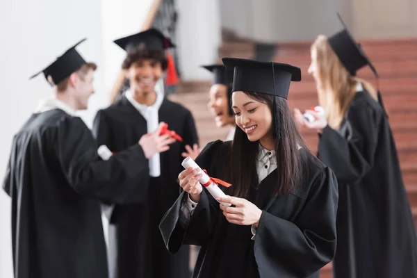 Sonriente asiático soltero mirando diploma en la universidad - foto de stock