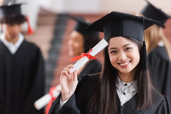 Asian graduate in cap smiling and holding diploma — Stock Photo