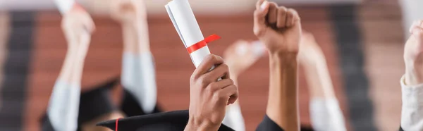 Vista recortada de estudiantes multiétnicos con diploma cerca de manos de amigos borrosos, pancarta - foto de stock