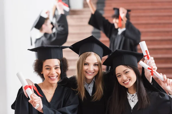 Souriant célibataire regardant la caméra près des amis interraciaux en robes académiques — Photo de stock